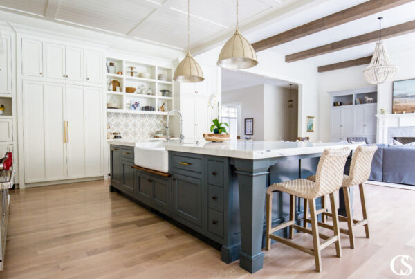 A spacious kitchen with a mix of white and gray cabinetry, a large central island, and pendant lighting. The room features wooden beams, a patterned backsplash, and a cozy seating area