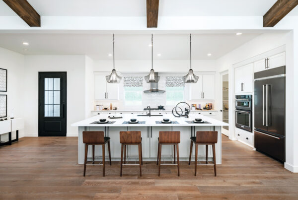 A spacious kitchen with white cabinets, a large central island, and pendant lighting over the island. The room features exposed wooden beams and a black framed glass door