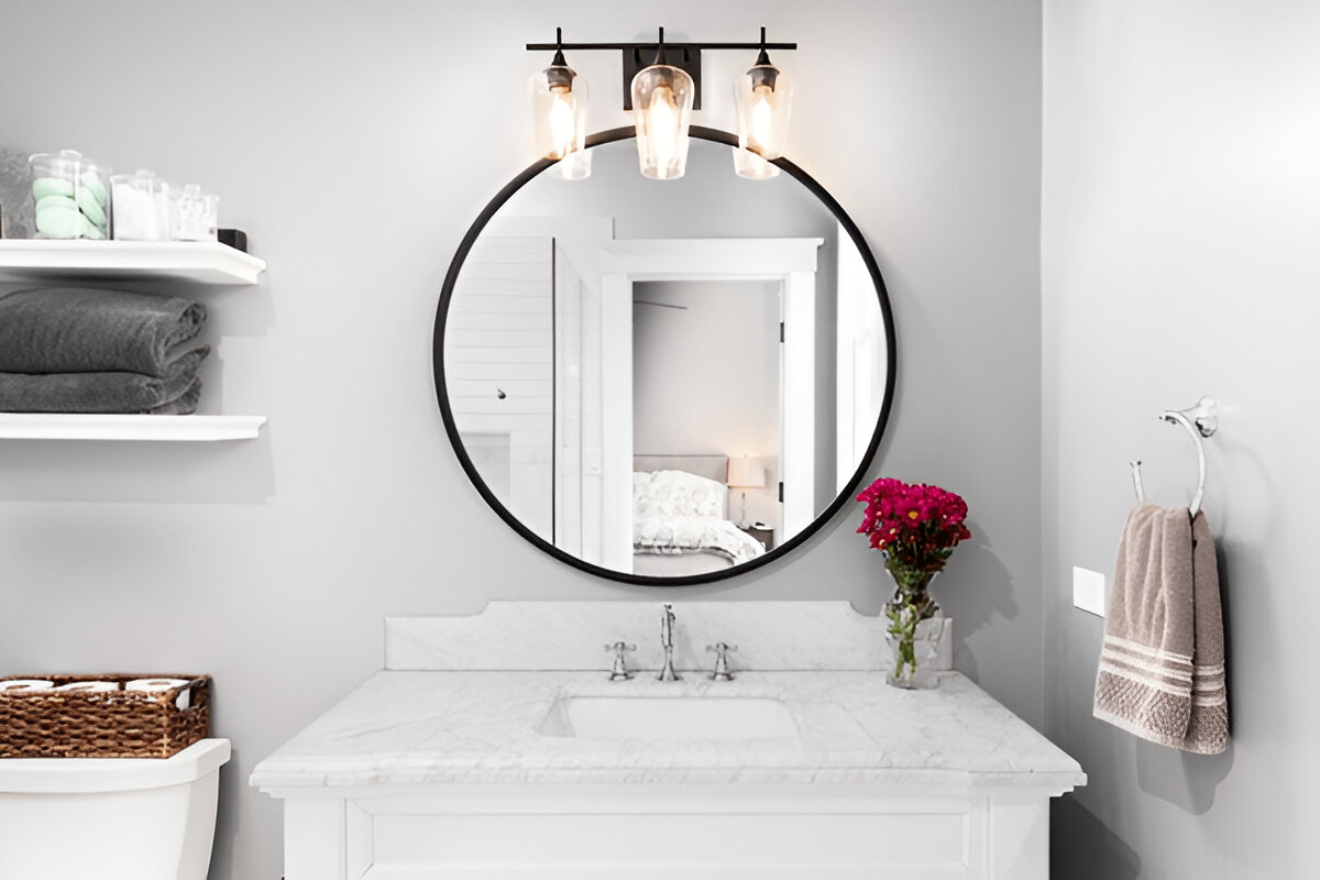 Modern bathroom with a white vanity, marble countertop, black-framed round mirror, industrial lighting, and open shelving with neatly folded towels