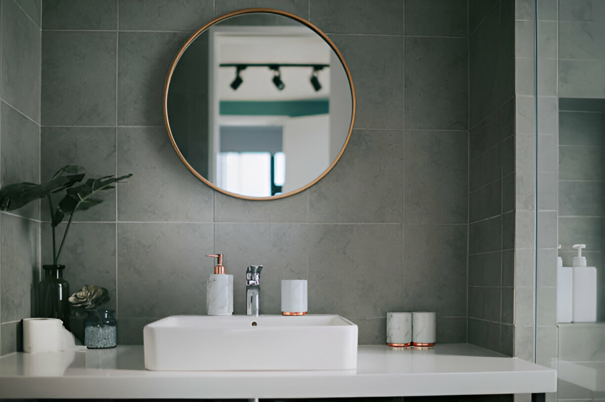 Bathroom interior with round gold-framed mirror, gray wall tiles, white sink on minimalist vanity, elegant accessories, and green plant decor