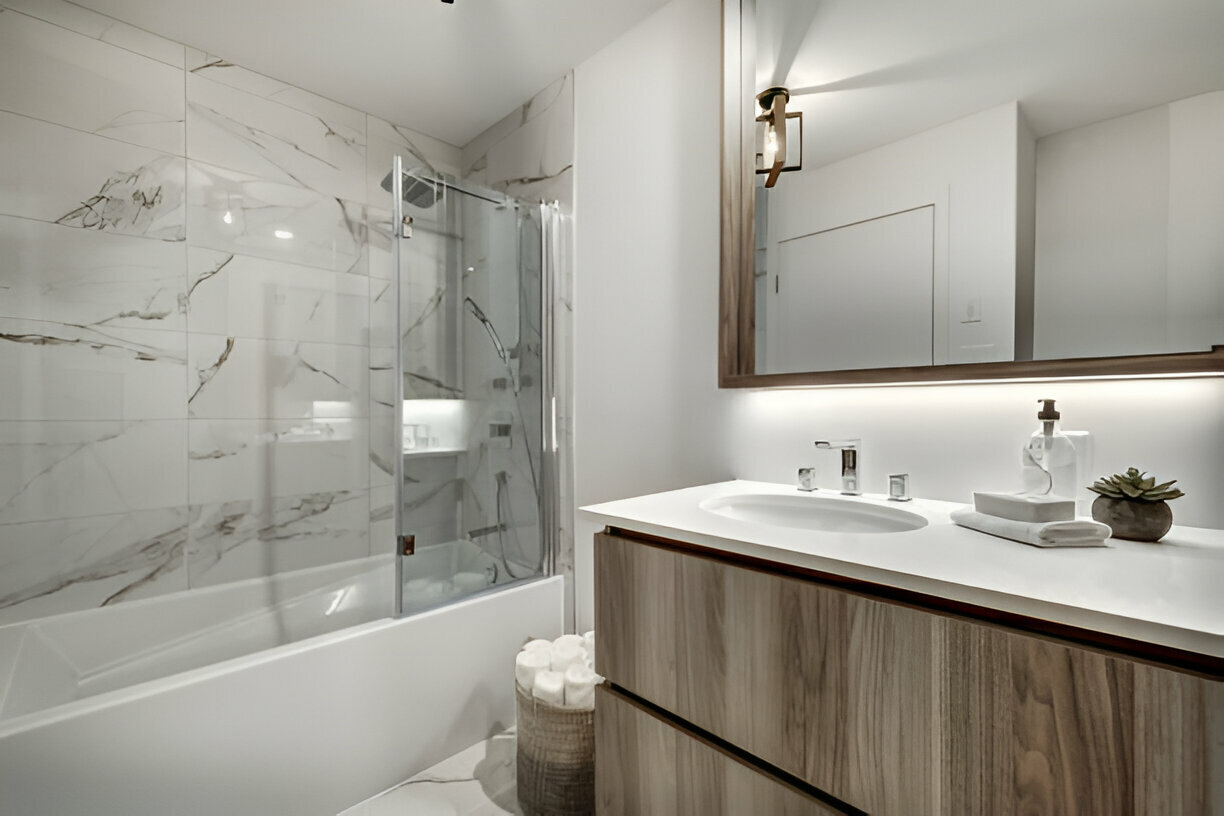 Floating wood vanity with white countertop, backlit mirror, marble tile walls, and a glass shower enclosure in a modern bathroom