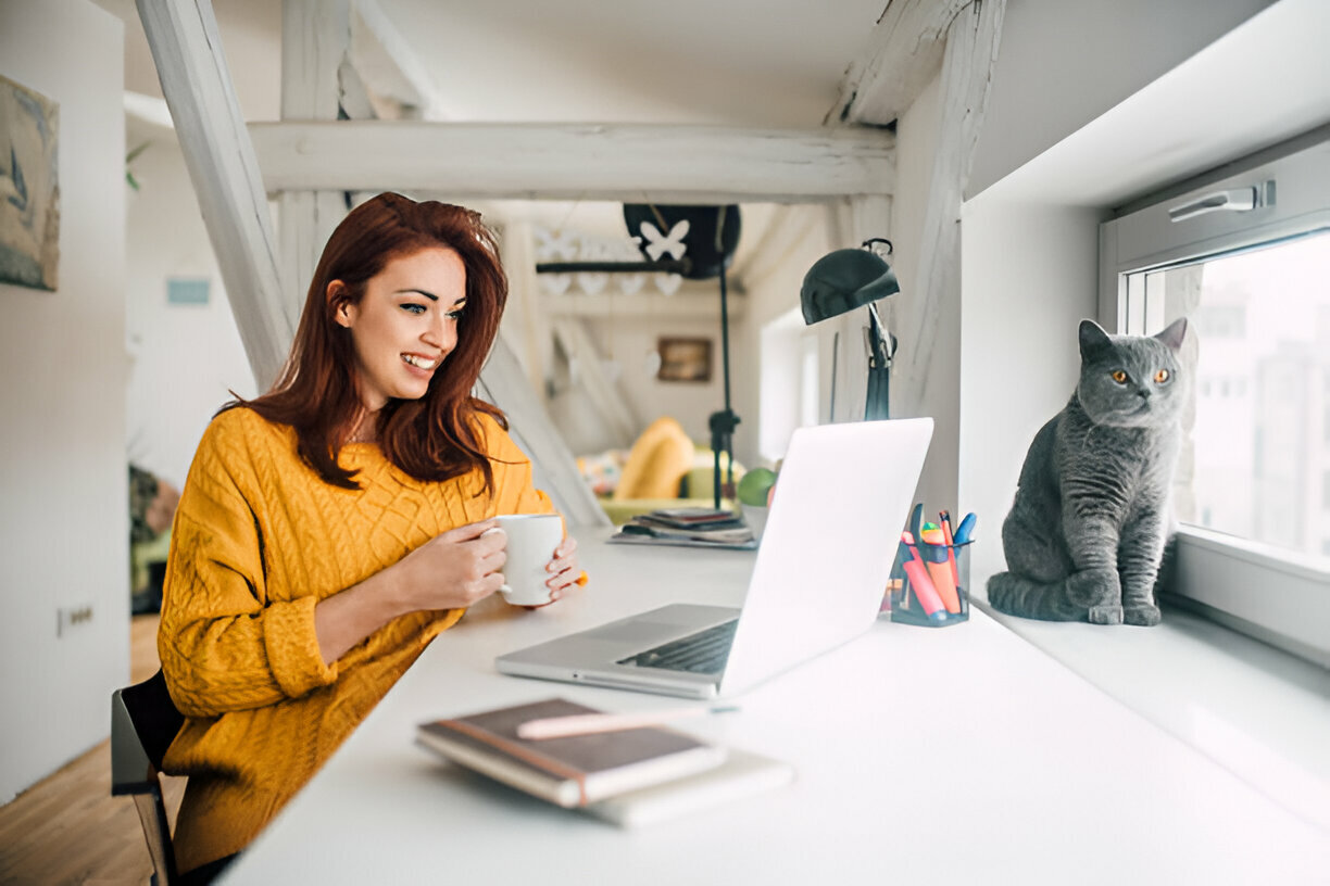 Woman in yellow sweater working on a laptop at home with a gray cat by the window