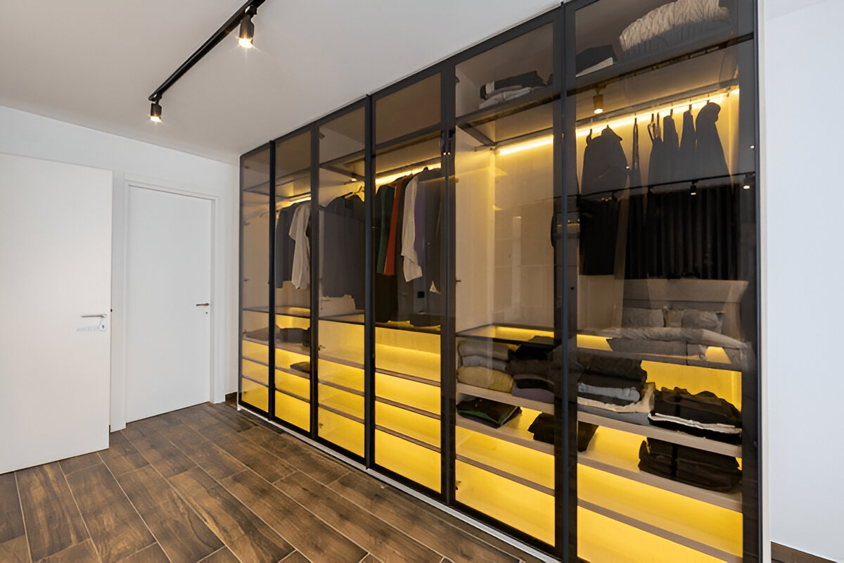 Modern glass wardrobe with internal yellow LED lighting and organized clothing storage.