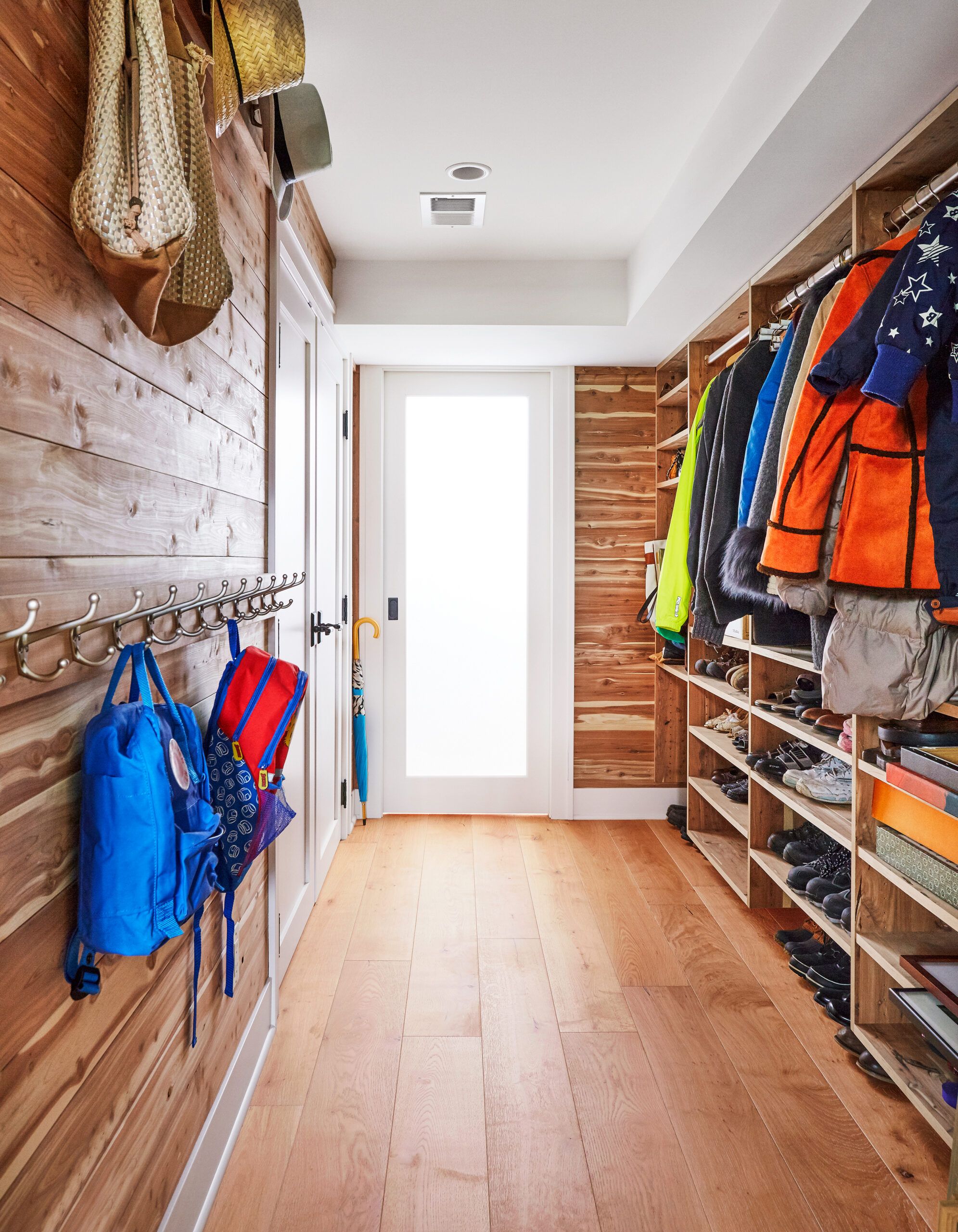 Mudroom with cedar plank walls, metal wall hooks holding backpacks, built-in open shelving with coats and shoe storage