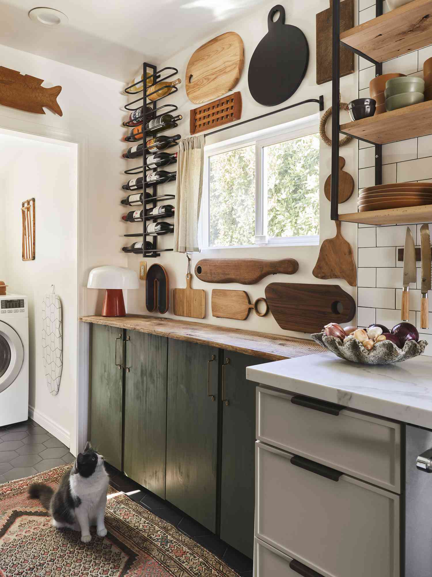 Eclectic kitchen with dark green lower cabinets, open wood shelves, mounted wine rack, wall-mounted cutting boards, and a black-and-white cat on a Persian-style rug.