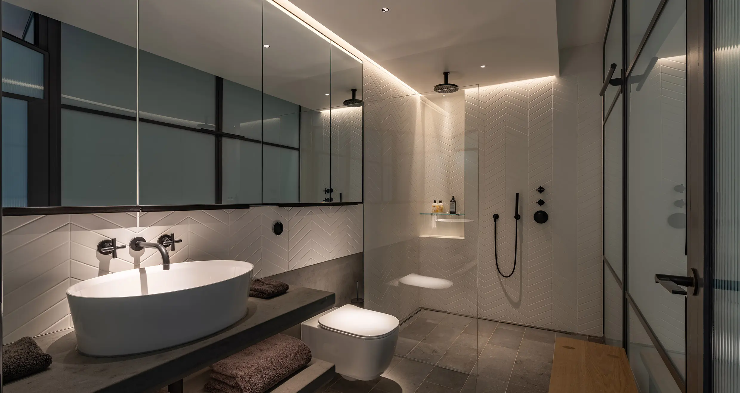 Modern minimalist bathroom featuring a floating vanity, vessel sink, LED strip lighting, matte black wall-mounted fixtures, walk-in shower, and herringbone tile walls