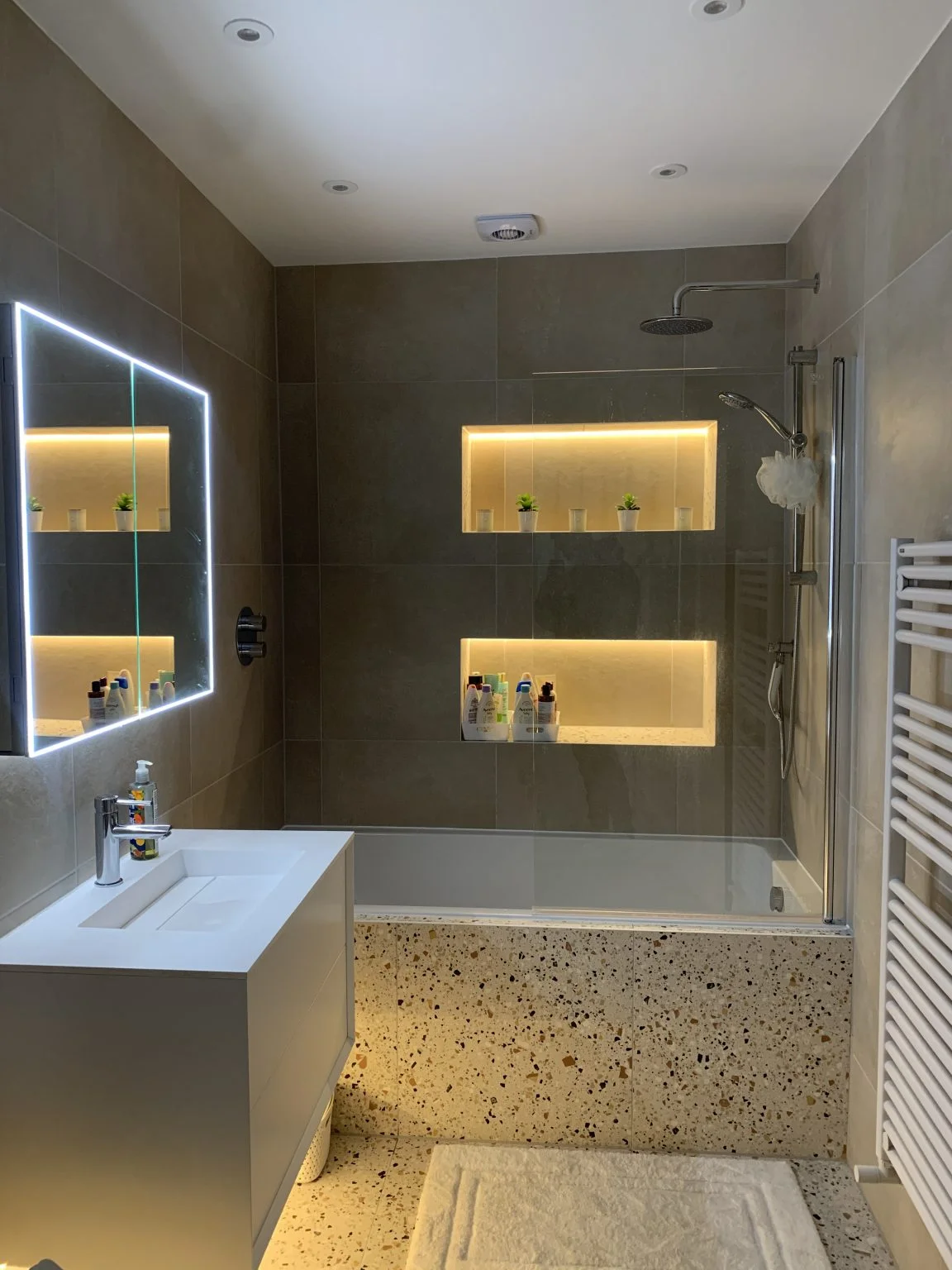 Modern bathroom featuring terrazzo bathtub paneling, LED-lit mirror, wall-mounted vanity, walk-in shower, and recessed shelves with ambient lighting