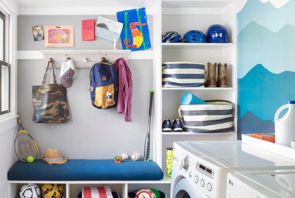 Colorful mudroom and laundry room combo with a built-in bench, wall hooks, cubbies for shoes and storage bins, and a painted blue mountain mural. Features include kids’ backpacks, helmets, sports gear, and front-load washer and dryer.