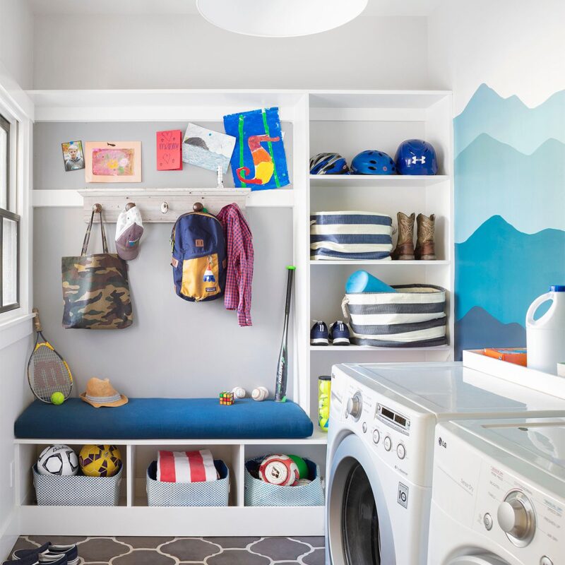 Colorful mudroom and laundry room combo with a built-in bench, wall hooks, cubbies for shoes and storage bins, and a painted blue mountain mural. Features include kids’ backpacks, helmets, sports gear, and front-load washer and dryer.