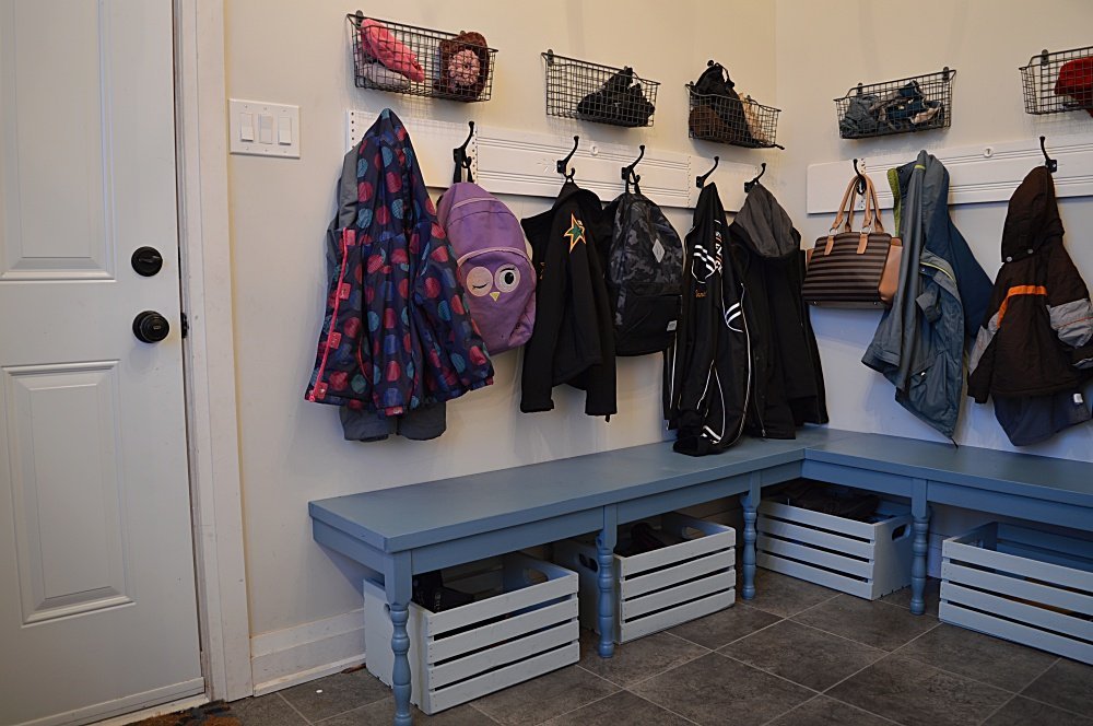 Cozy mudroom featuring corner blue-painted wooden benches with white crate storage underneath, wall hooks holding coats and backpacks, and wire baskets mounted above for hats and gloves.