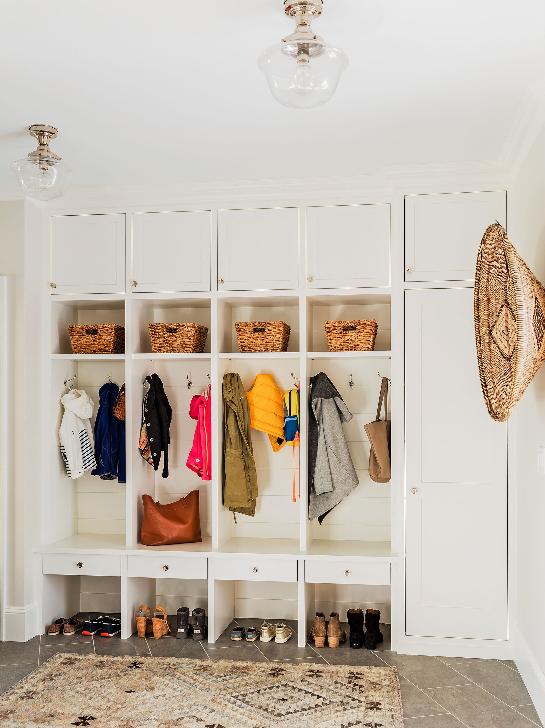 Light-filled mudroom featuring custom white built-in cubbies with upper cabinets and wicker baskets, hooks for jackets and bags, drawers, and lower open shelves for shoes.