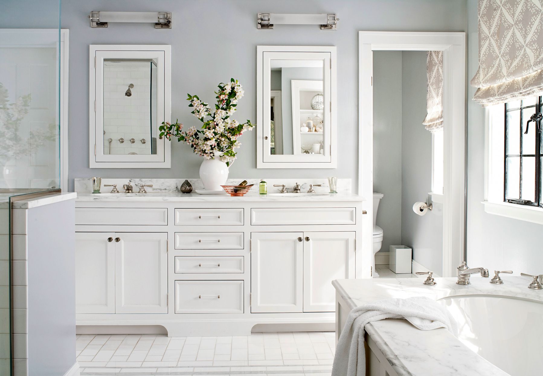 Traditional white bathroom with dual sink vanity, marble countertops, built-in mirrors, chrome fixtures, and fresh flower arrangement