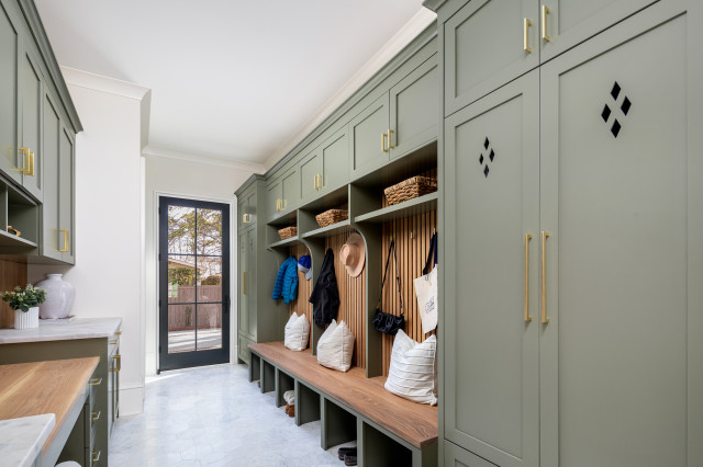 Spacious mudroom with sage green built-in cabinets, wood bench seating, upper storage baskets, gold hardware, and vertical paneling. A glass door at the end lets in natural light.