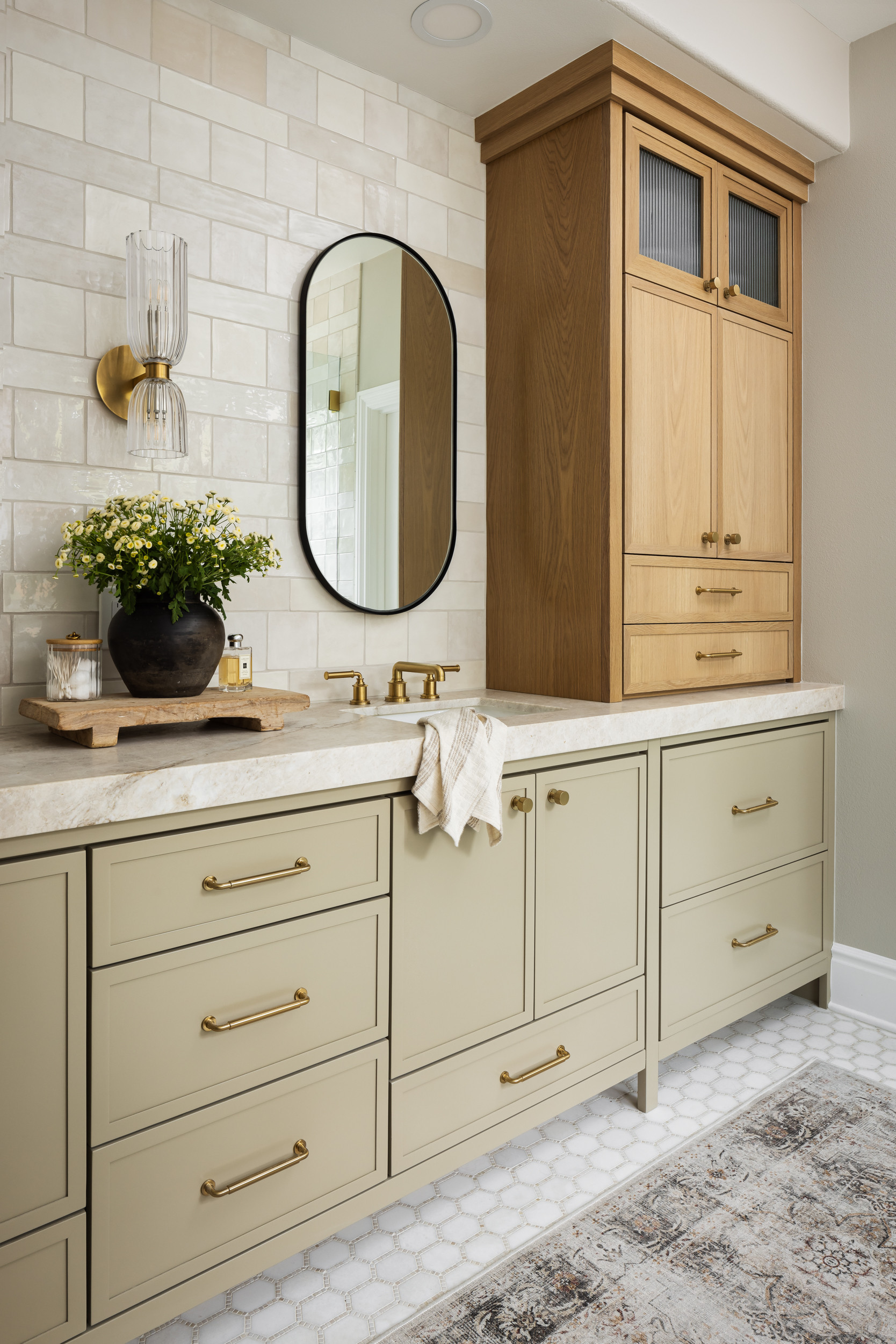 Bathroom with sage green cabinets, gold hardware, wood tower cabinet, marble countertop, oval mirror, and wall-mounted glass sconce