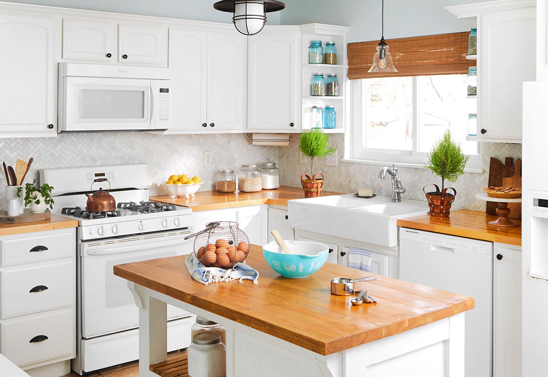 Airy white kitchen with natural wood countertops, a central island, open shelving, and a farmhouse sink under a window.
