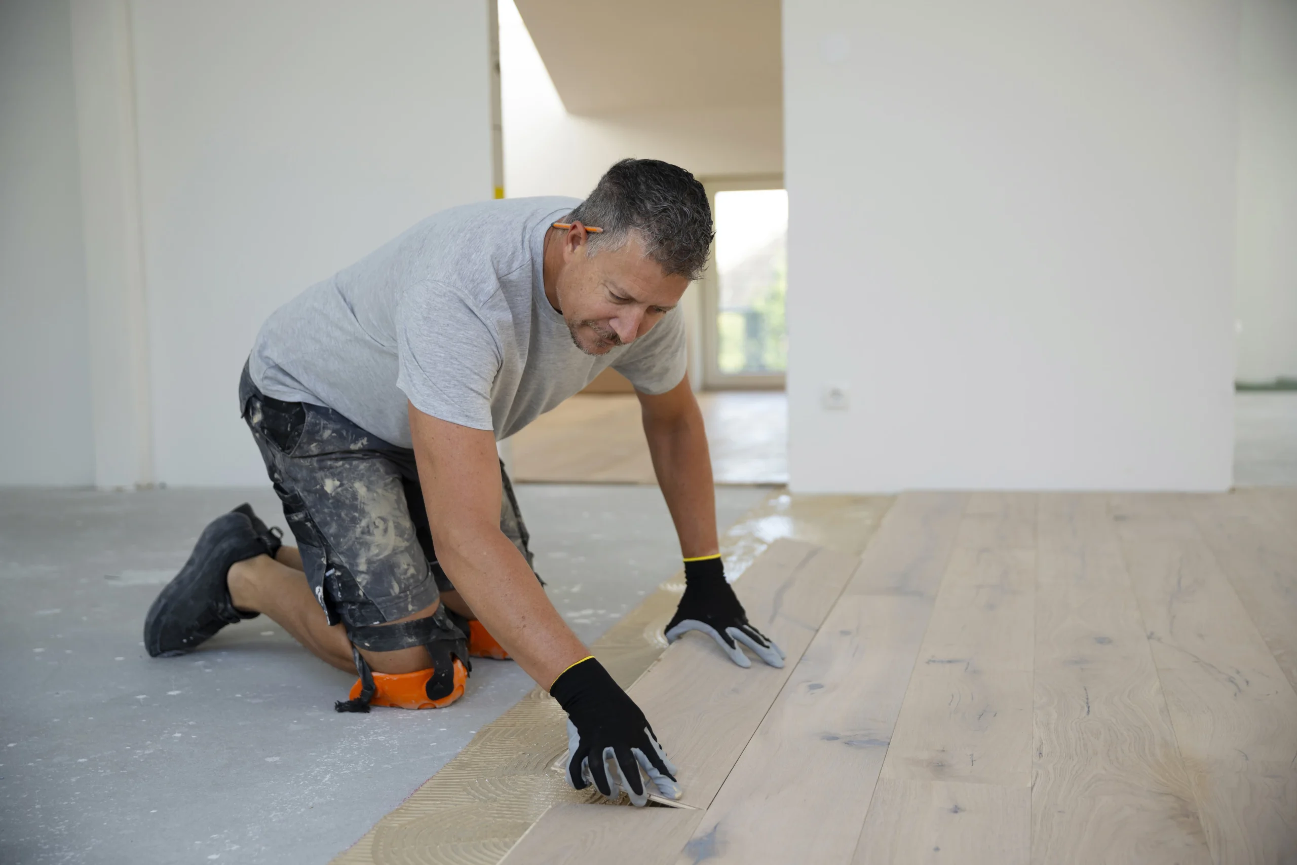 Worker installing light timber flooring on a concrete subfloor, using adhesive and alignment tools.