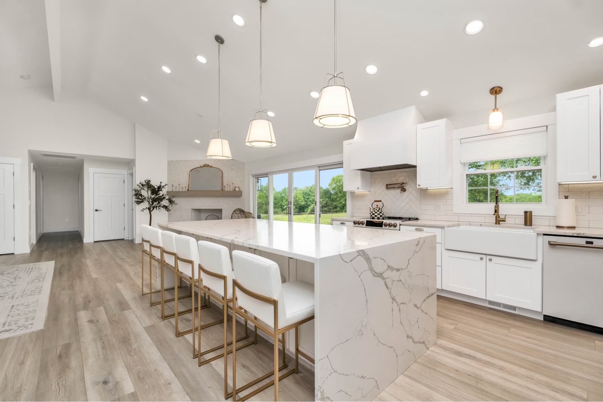  Open-concept white kitchen featuring a waterfall marble island, white shaker cabinets, gold-accented stools, and a large farmhouse sink.