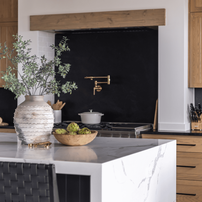 Kitchen featuring light natural wood cabinets, a white marble waterfall island, black countertops and backsplash, and brass pot filler.