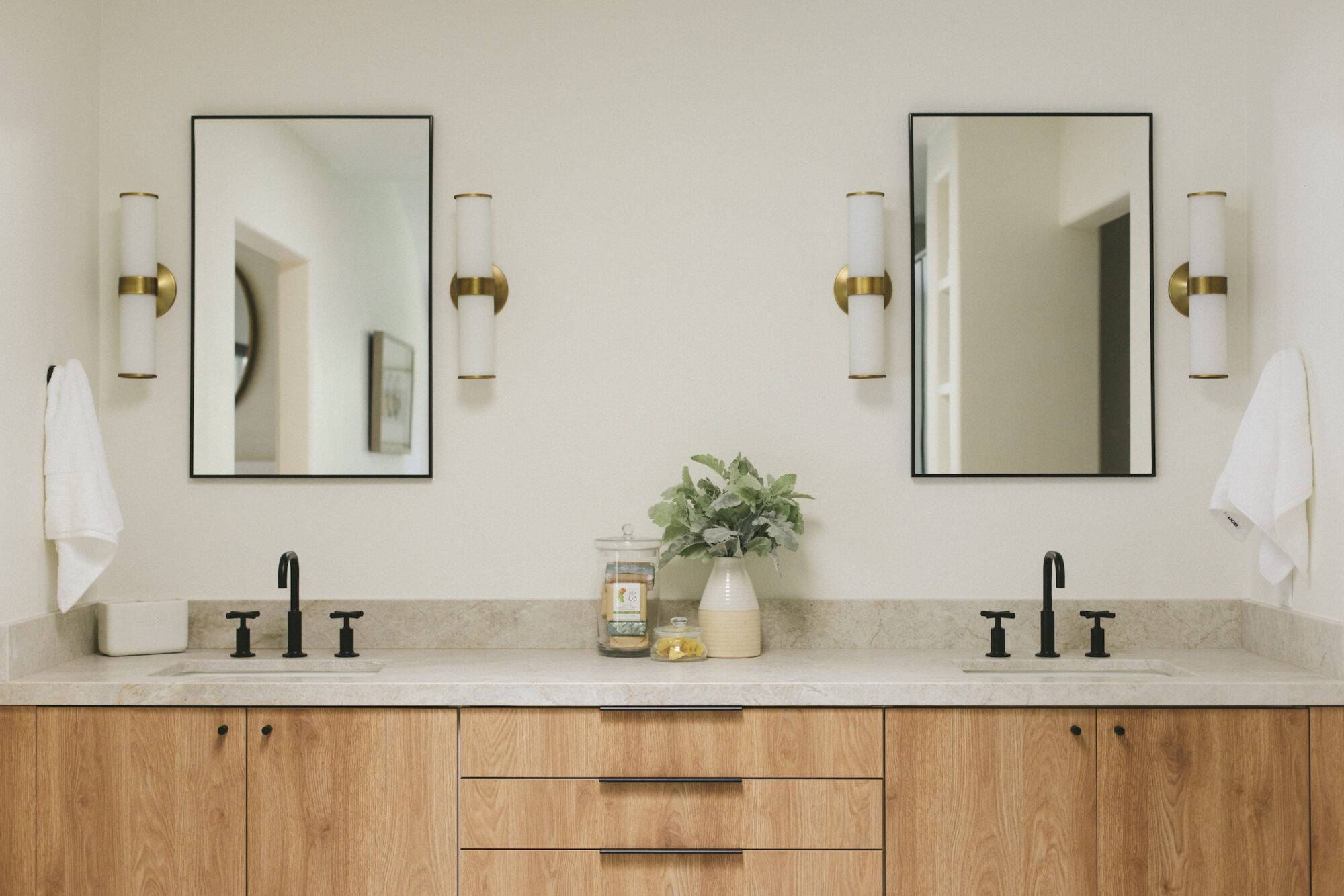  Bathroom with dual sinks, light wood cabinetry, black faucets, beige stone countertop, and twin mirrors with brass sconces.