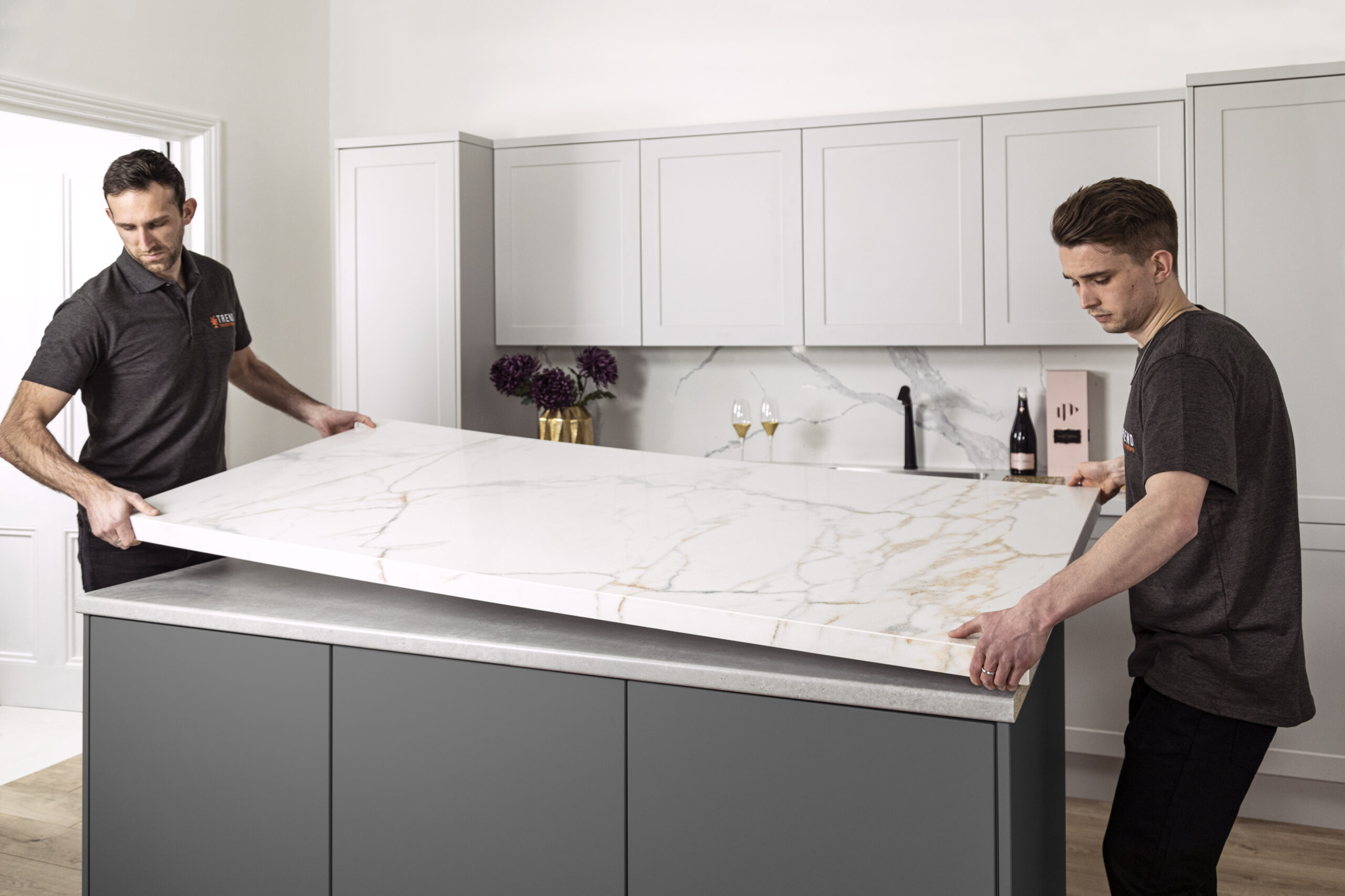 Two workers installing a white marble countertop with grey and gold veining onto a dark grey kitchen island.