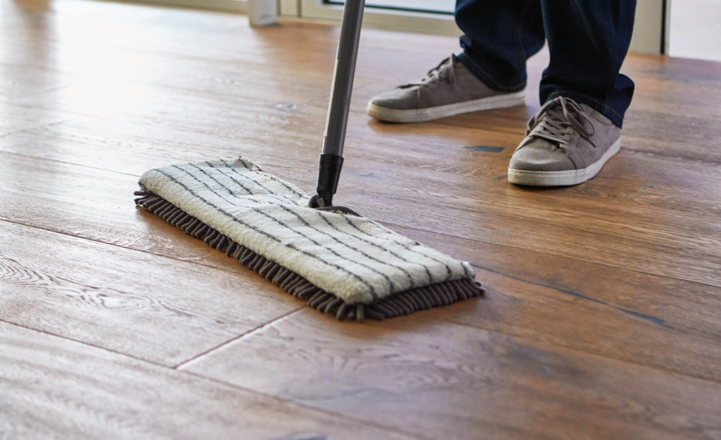 Person using a microfiber mop to clean wood-look laminate flooring, wearing grey sneakers and dark jeans.