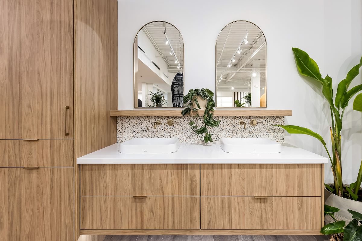  Double bathroom vanity with light wood cabinets, terrazzo backsplash, dual white vessel sinks, gold fixtures, and arched mirrors.