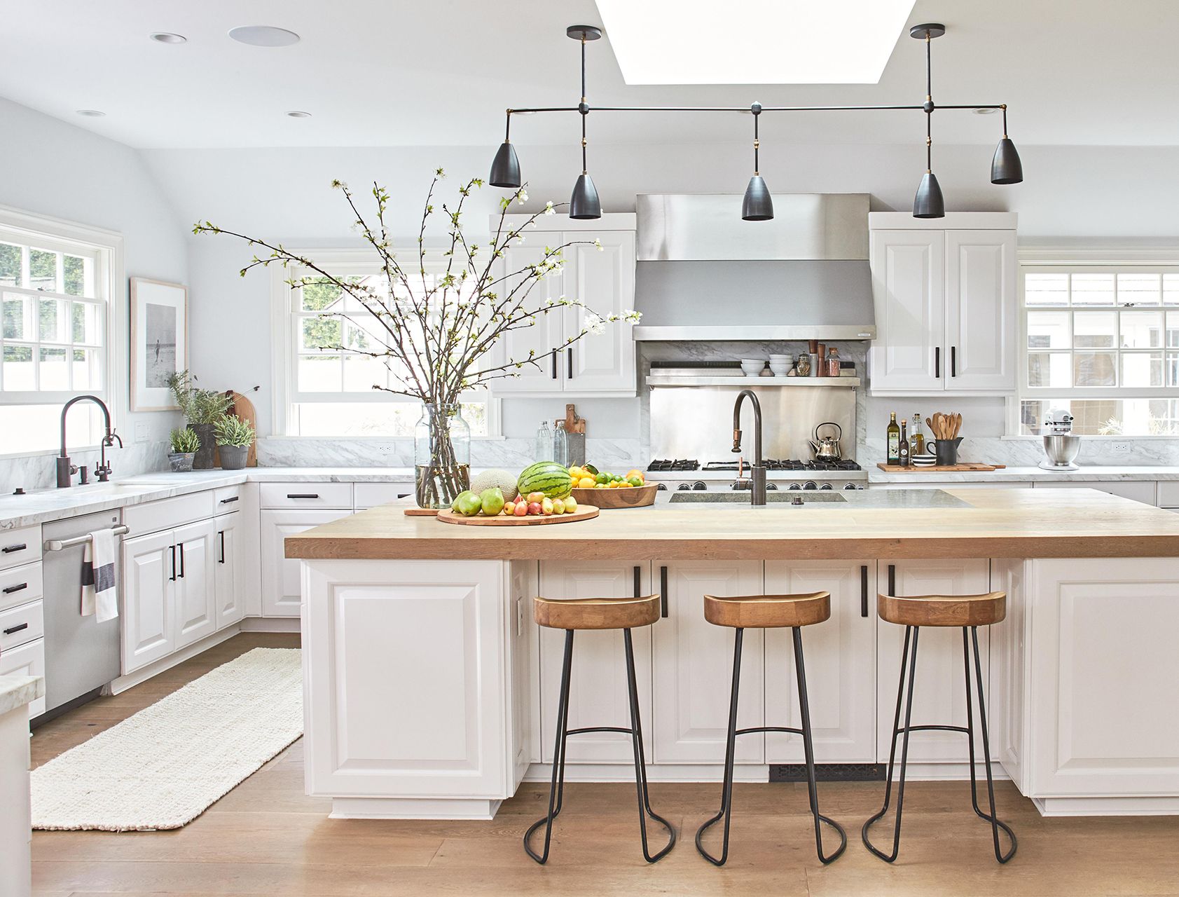 Spacious white kitchen featuring a large island with wood countertop, black metal stools, matte black fixtures, and stainless steel appliances.