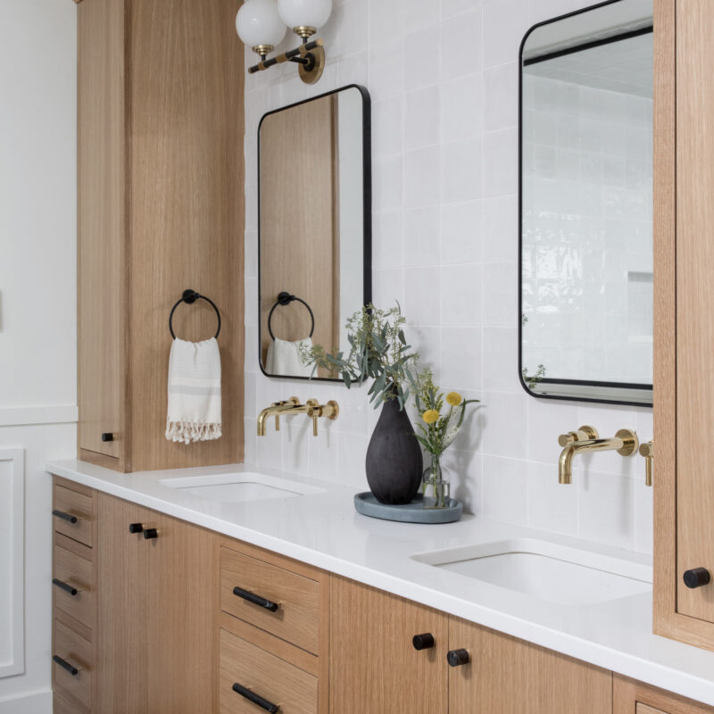 Modern bathroom with natural wood cabinetry, dual sinks, and wall-mounted brass faucets.