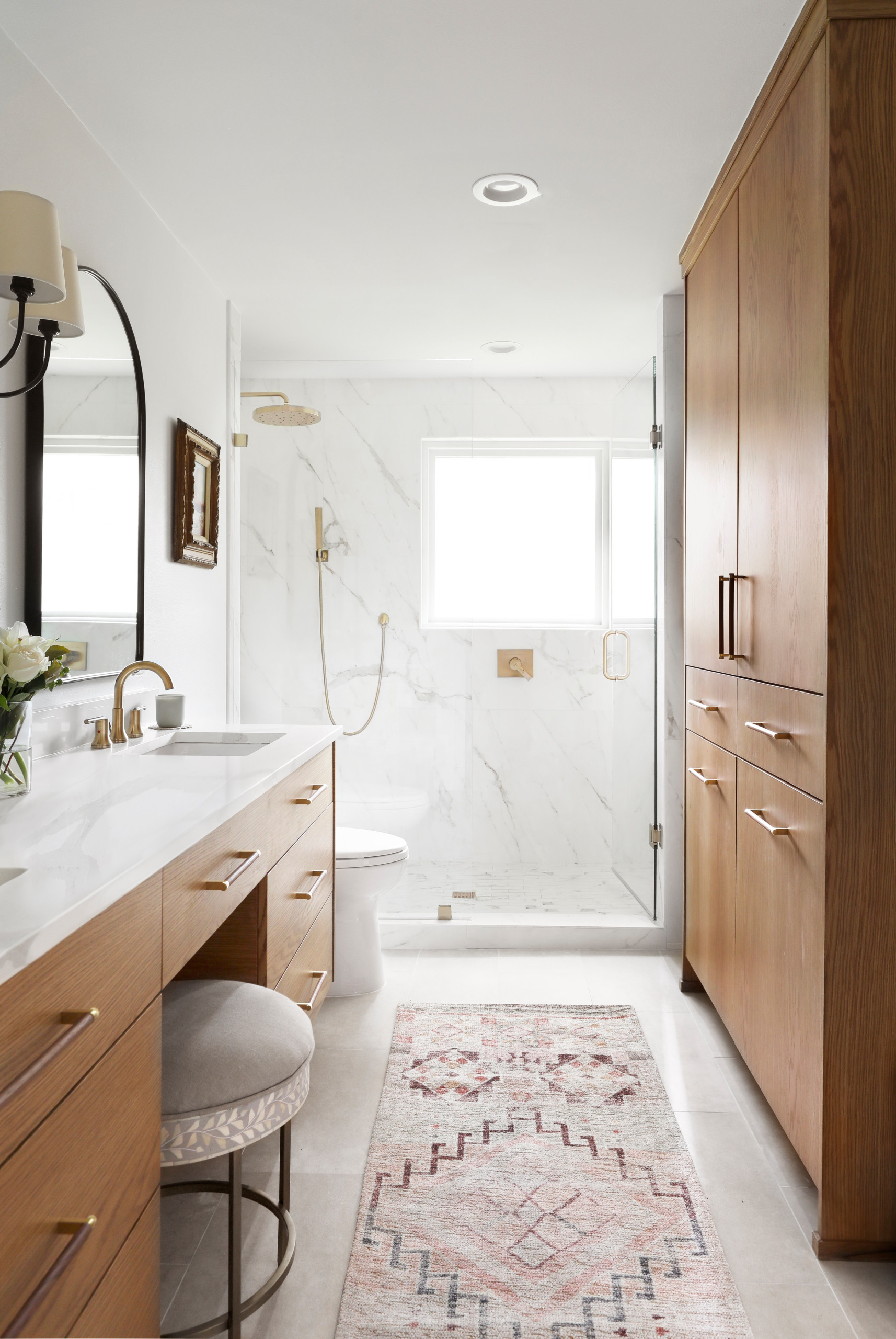 Bright modern bathroom with wooden vanity, marble shower, and patterned rug.