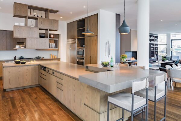 A contemporary kitchen with light wood cabinetry, sleek gray countertops, and modern pendant lighting. Open shelving adds functionality and style, while the space flows seamlessly into a cozy living area with a fireplace.