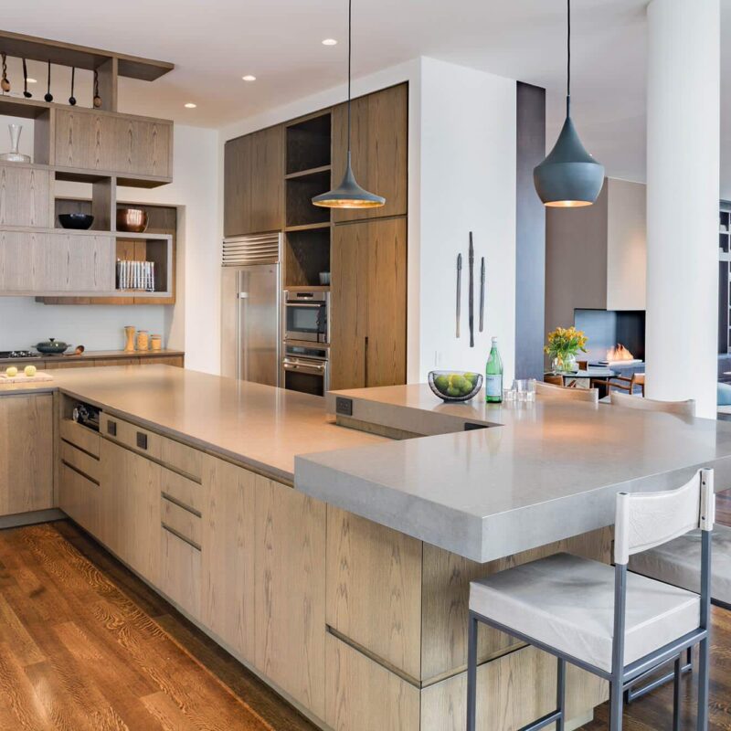 A contemporary kitchen with light wood cabinetry, sleek gray countertops, and modern pendant lighting. Open shelving adds functionality and style, while the space flows seamlessly into a cozy living area with a fireplace.