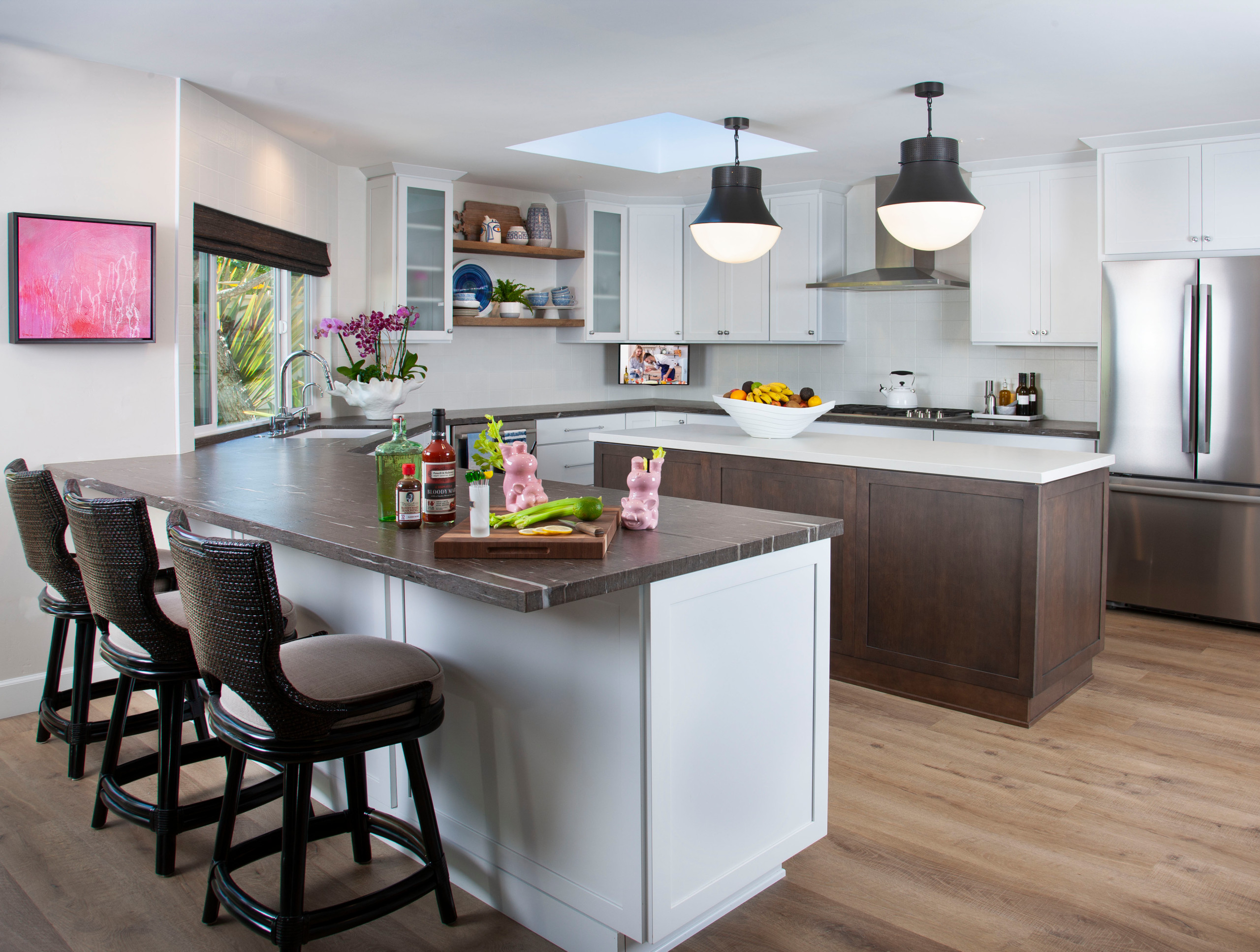A modern kitchen with an open layout featuring a large island, white cabinetry, wooden accents, and sleek stainless steel appliances. The space is illuminated by black pendant lights and a skylight, with a stylish mix of decor and functional elements.