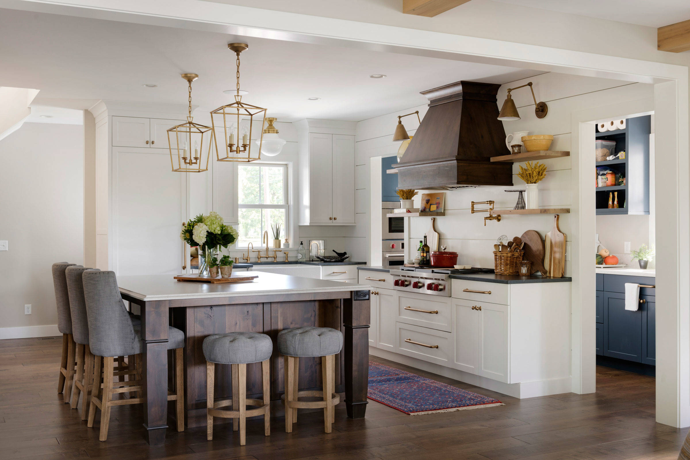 A transitional farmhouse kitchen featuring a large central island with grey stools, brass light fixtures hanging above, and white cabinets with gold handles. There is a dark wood range hood and an open shelf with plants and kitchen items on the wall.