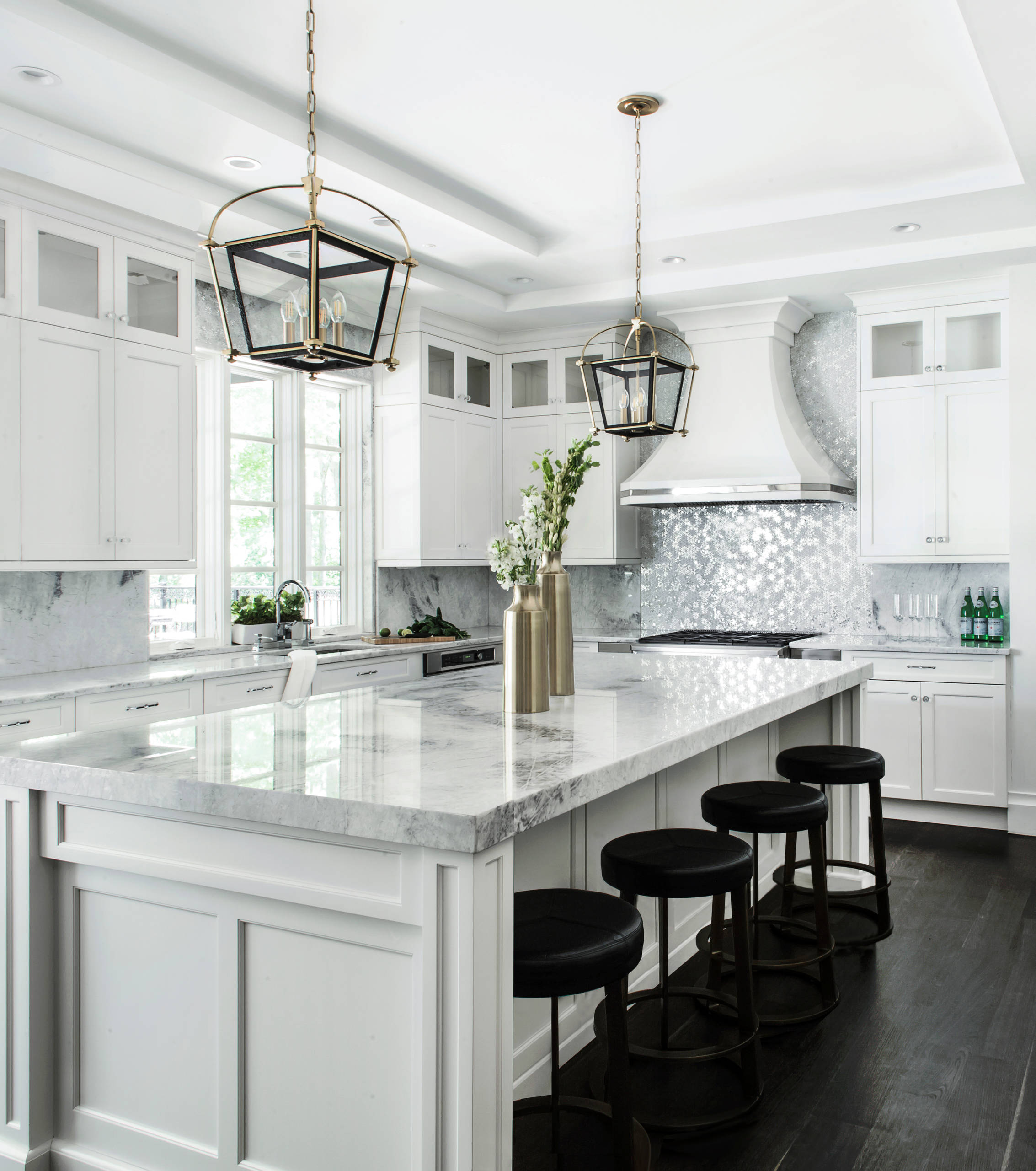 A luxurious white kitchen featuring a large marble island, elegant black stools, and sophisticated lantern-style pendant lighting. The backsplash is adorned with shimmering mosaic tiles, and the space is complemented by dark wooden flooring.