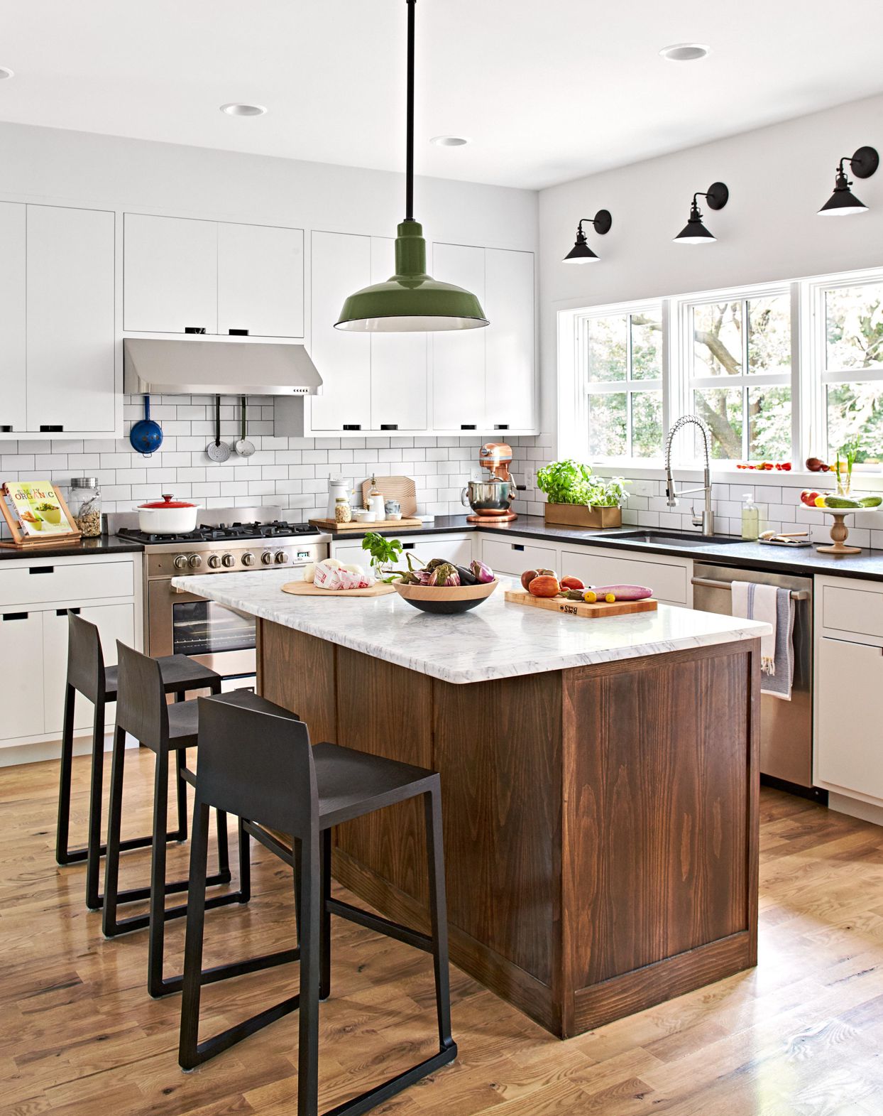 A bright kitchen with white cabinetry, subway tile backsplash, and a central island featuring a wooden base and marble countertop. Black bar stools and a green pendant light add contrasting accents, while natural light streams through large windows.